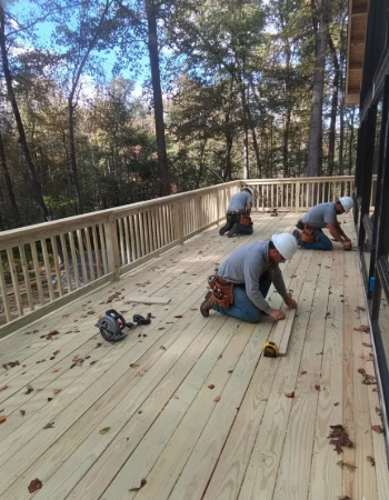 Three construction workers are kneeling on a large wooden deck in a wooded area, installing the final deck boards