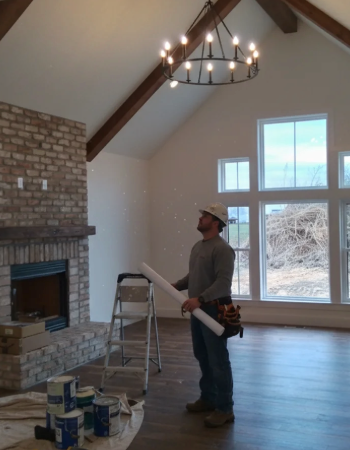 A worker surveys a finished living room with a brick fireplace and vaulted ceiling with wooden beams