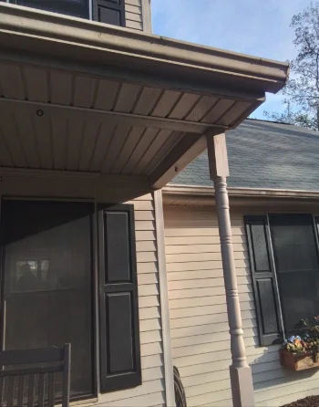 A beige vinyl-sided house features dark shutters and a covered porch supported by a single turned post